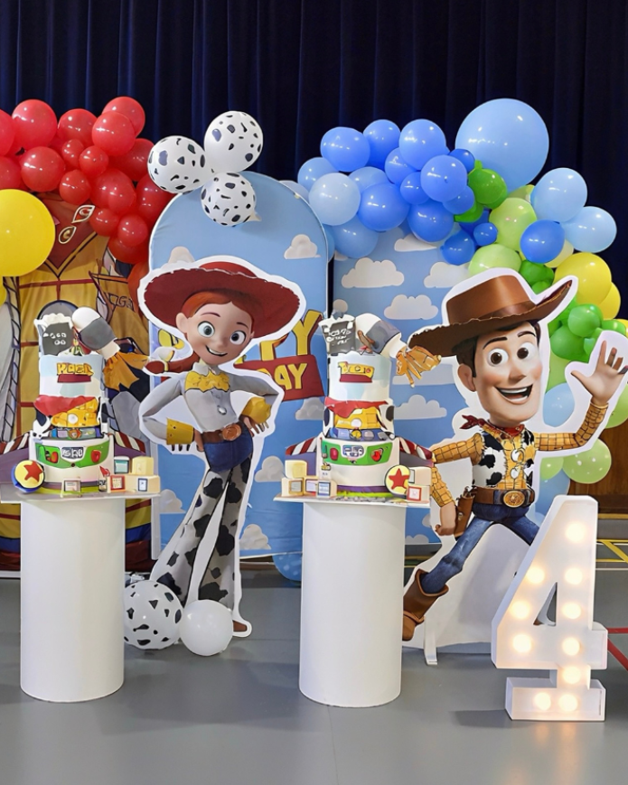 A children's birthday party setup featuring Woody and Buzz Lightyear cutout figures on white pedestals, decorated with blue, red, yellow, and green balloon clusters, a blue sky backdrop, and large colorful letter blocks spelling part of 'SIXTY'.