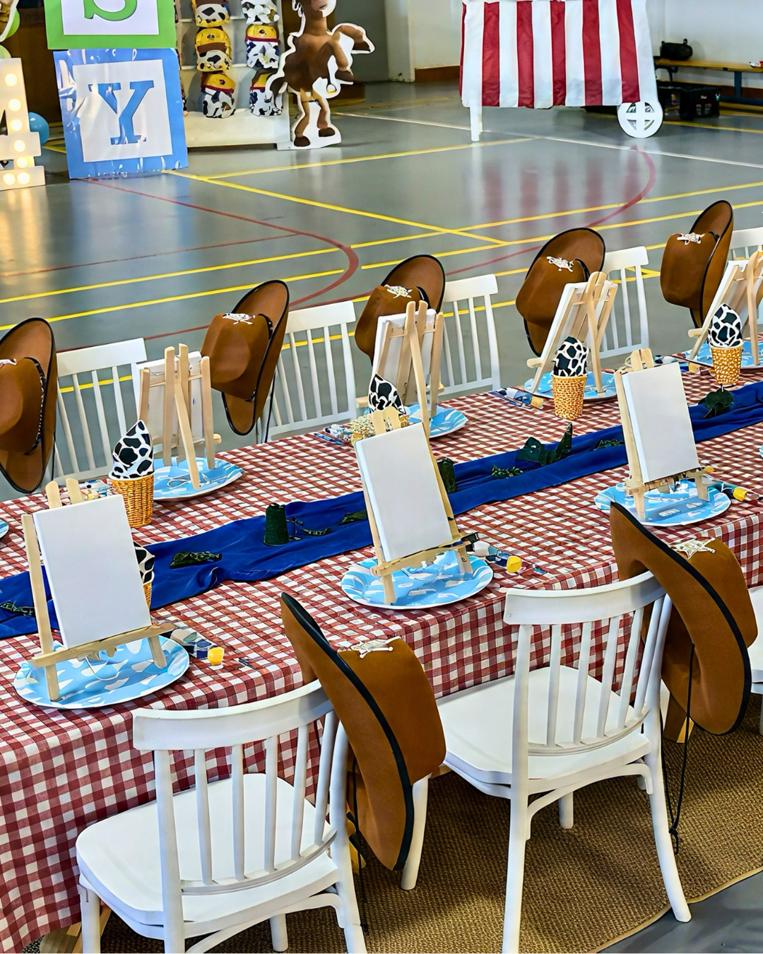A long table set for a western-themed party, featuring red and white checkered tablecloths, white chairs with brown cowboy hats on the backs, blue napkins, and small easels with painted canvases.