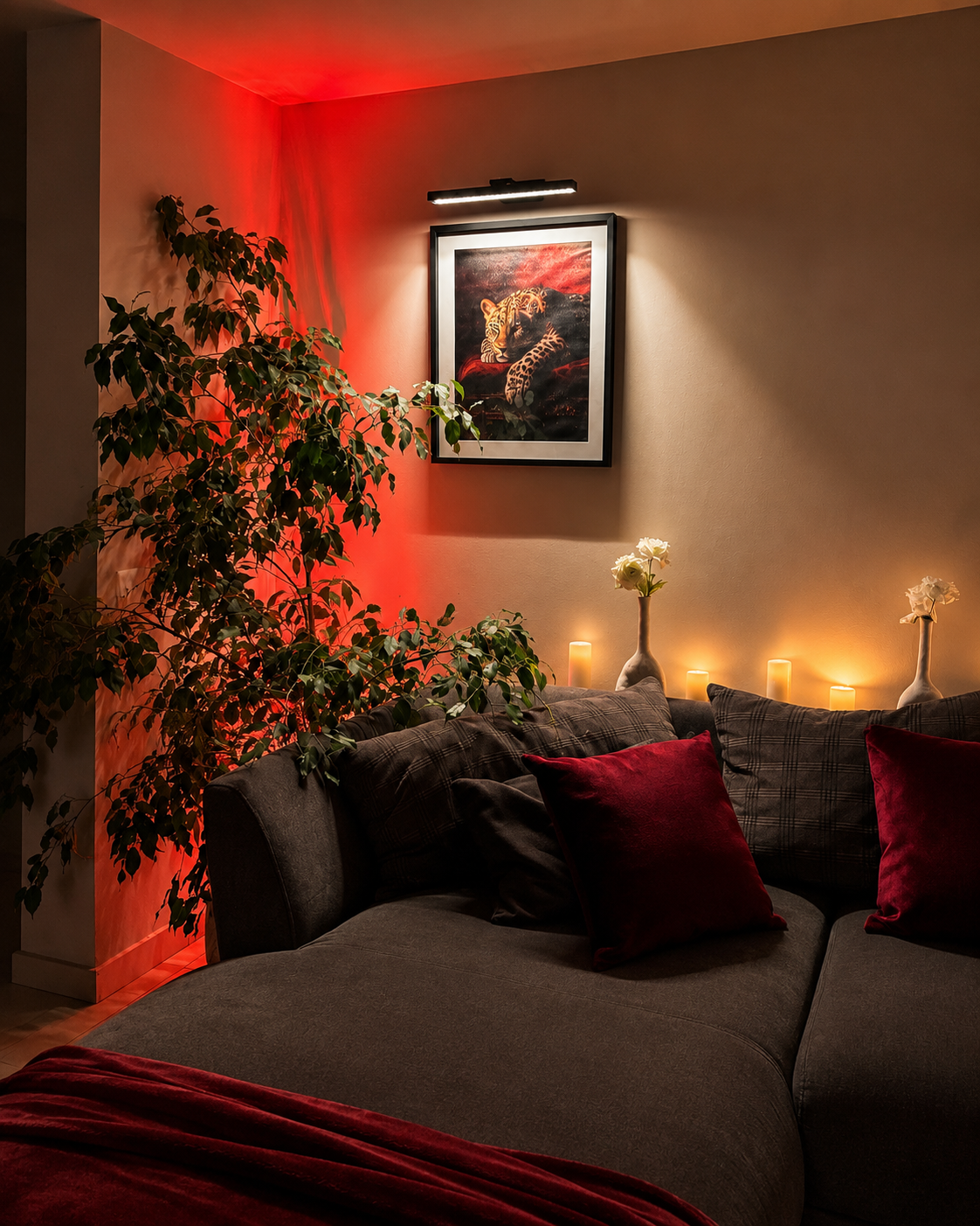 A dark gray sectional sofa in a cozy living room setting, styled with deep red throw pillows and a red blanket. A framed art print hangs on the beige wall above the sofa, and warm candles and red ambient lighting create a warm atmosphere.