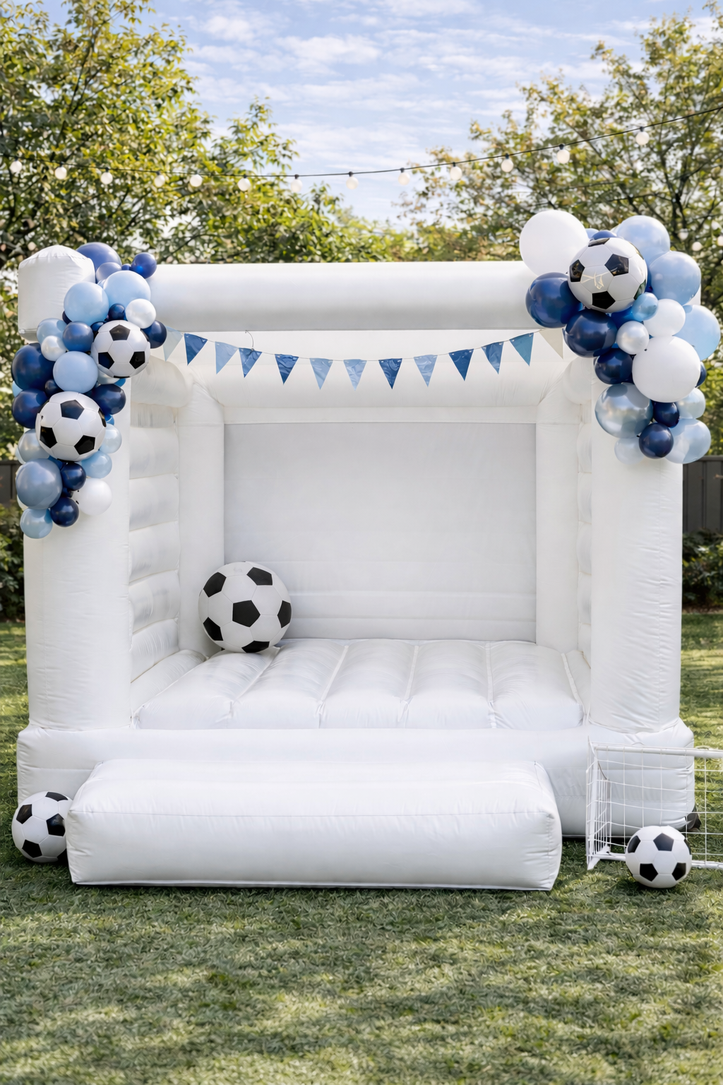 A white inflatable bounce house decorated with blue and white soccer-themed balloons, a blue bunting banner, and soccer balls, set up on a green lawn outdoors.