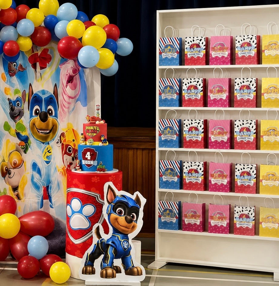 A Paw Patrol themed party setup featuring a large character backdrop, balloon garland, a tiered cake, a character cutout, and shelves of matching party bags.