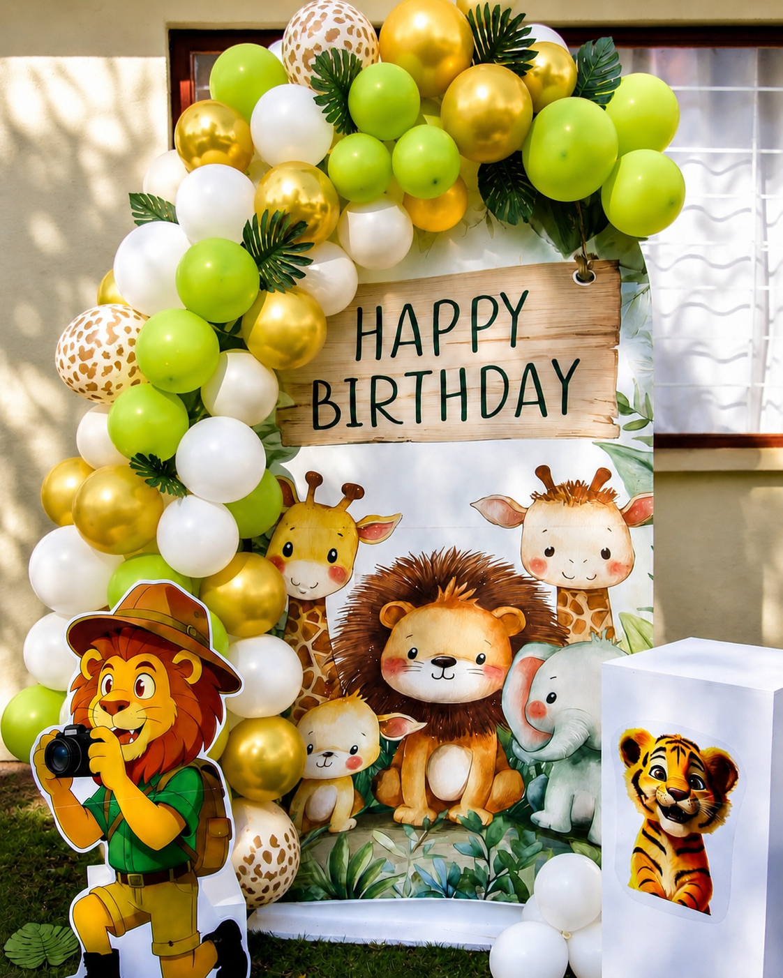 Safari-themed birthday party decoration set featuring a balloon garland arch in green, gold, and white, a 'HAPPY BIRTHDAY' sign, a lion cartoon cutout, and a matching white party popper box with a tiger illustration.