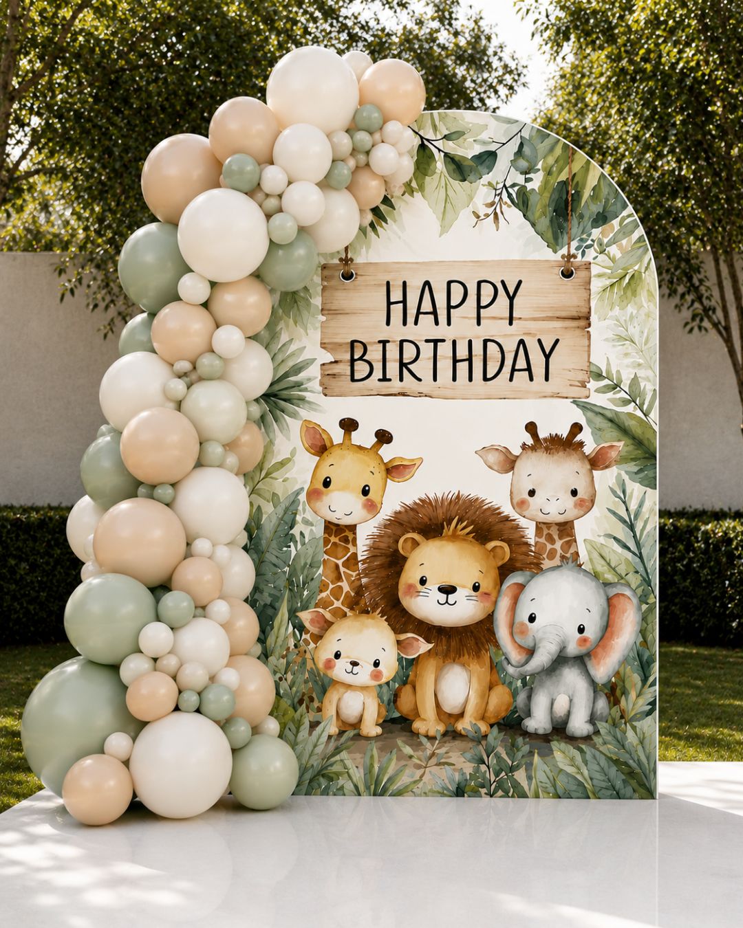 A safari-themed birthday backdrop stand featuring cartoon animals including giraffes, a lion, a baby elephant, and a baby hippo under a 'HAPPY BIRTHDAY' sign, styled with a matching balloon garland in an outdoor garden setting.