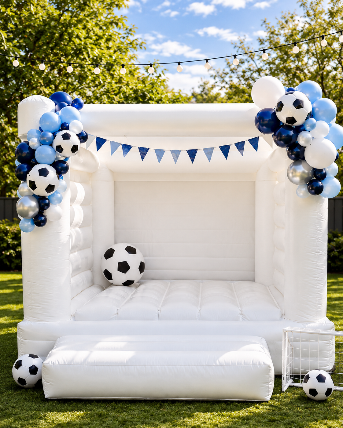 A white inflatable bouncy castle decorated for a soccer party, featuring blue and white balloon garlands with soccer balls, set on a green lawn outdoors.