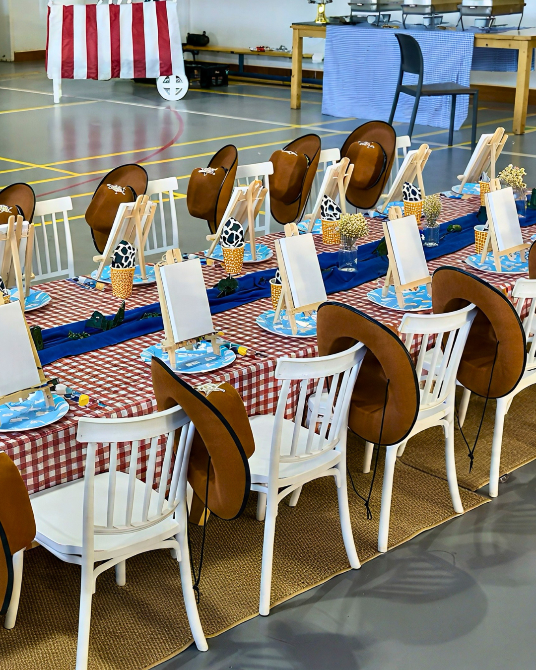 A long table set up for a themed event, featuring red and white checkered tablecloths, blue plates, small easels, and multiple brown cowboy hats draped over white chairs in a large indoor hall.