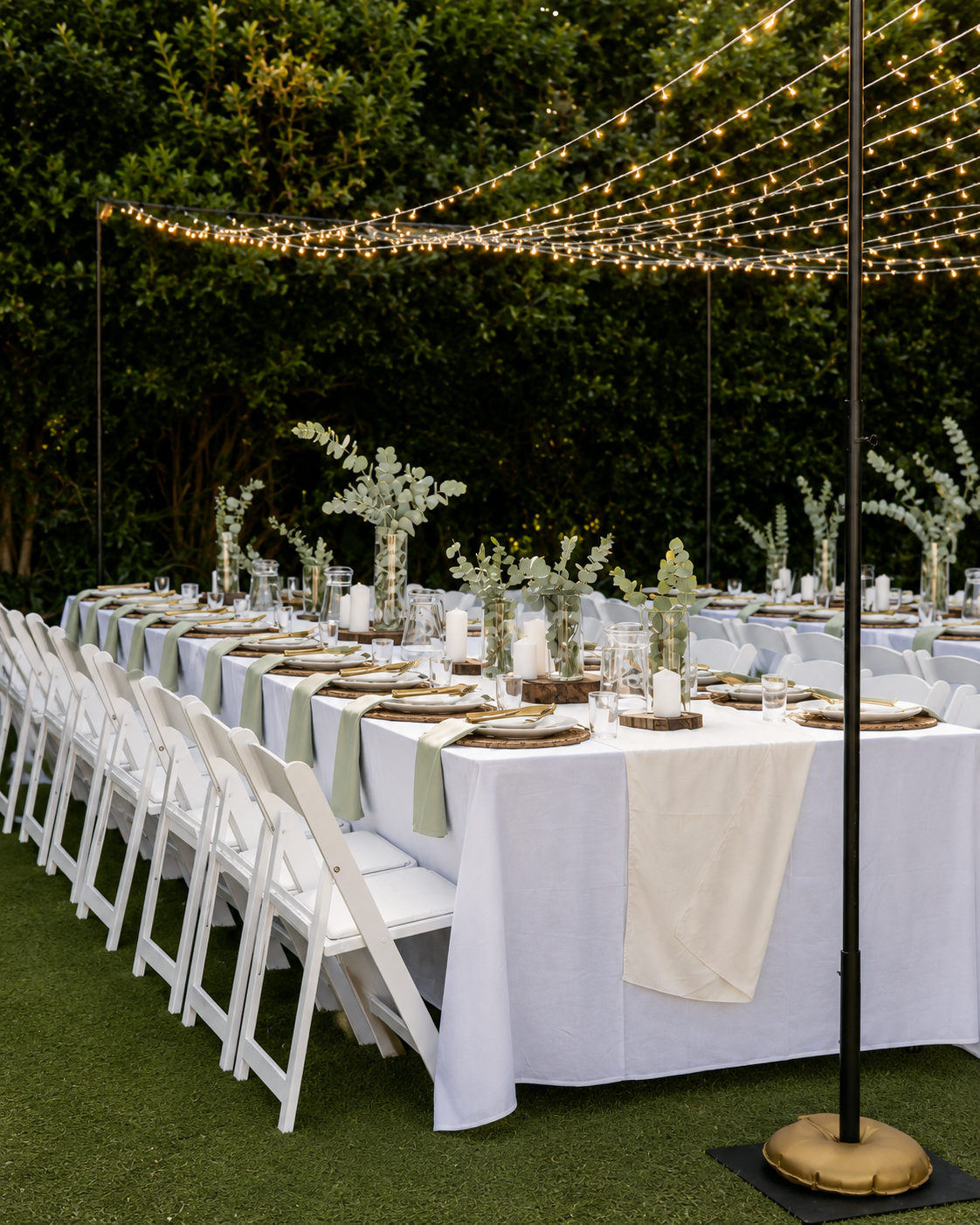 Outdoor wedding reception tables set on green grass under a black patio umbrella with warm string lights. The tables are covered in white tablecloths with white and sage green runners, white folding chairs, and floral centerpieces.
