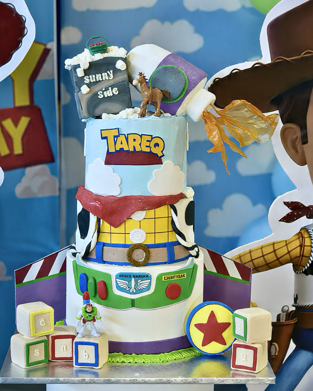A three-tiered custom cake decorated to look like Woody from Toy Story, with a Buzz Lightyear section on the bottom tier, toy-themed decorations, and a small cowboy figurine on top, set against a blue sky and clouds backdrop.