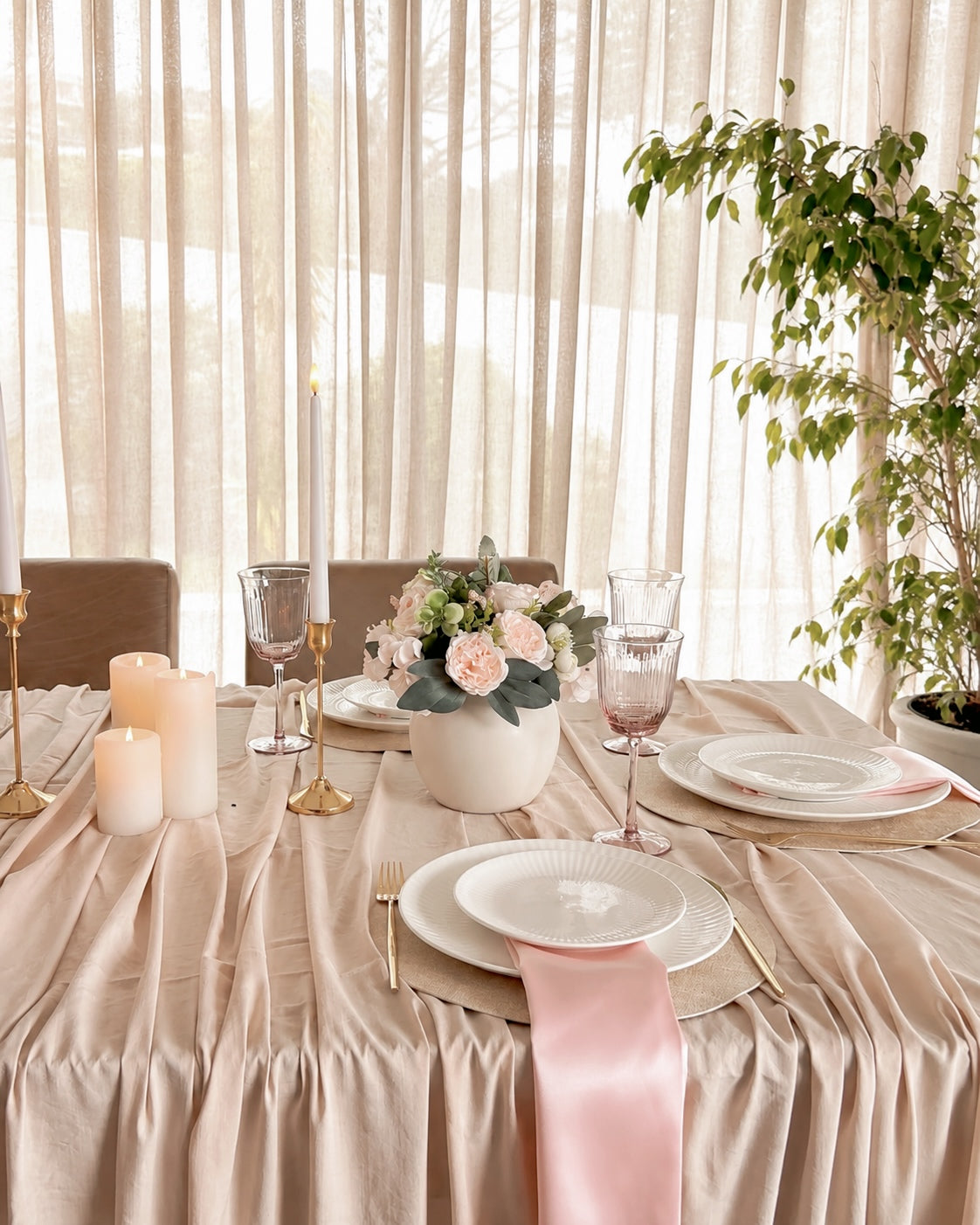 A beautifully styled dining table draped in a champagne beige tablecloth, set with white plates, gold cutlery, pink satin napkins, glassware, candles, and a floral centerpiece. The background features sheer curtains and a potted green plant.