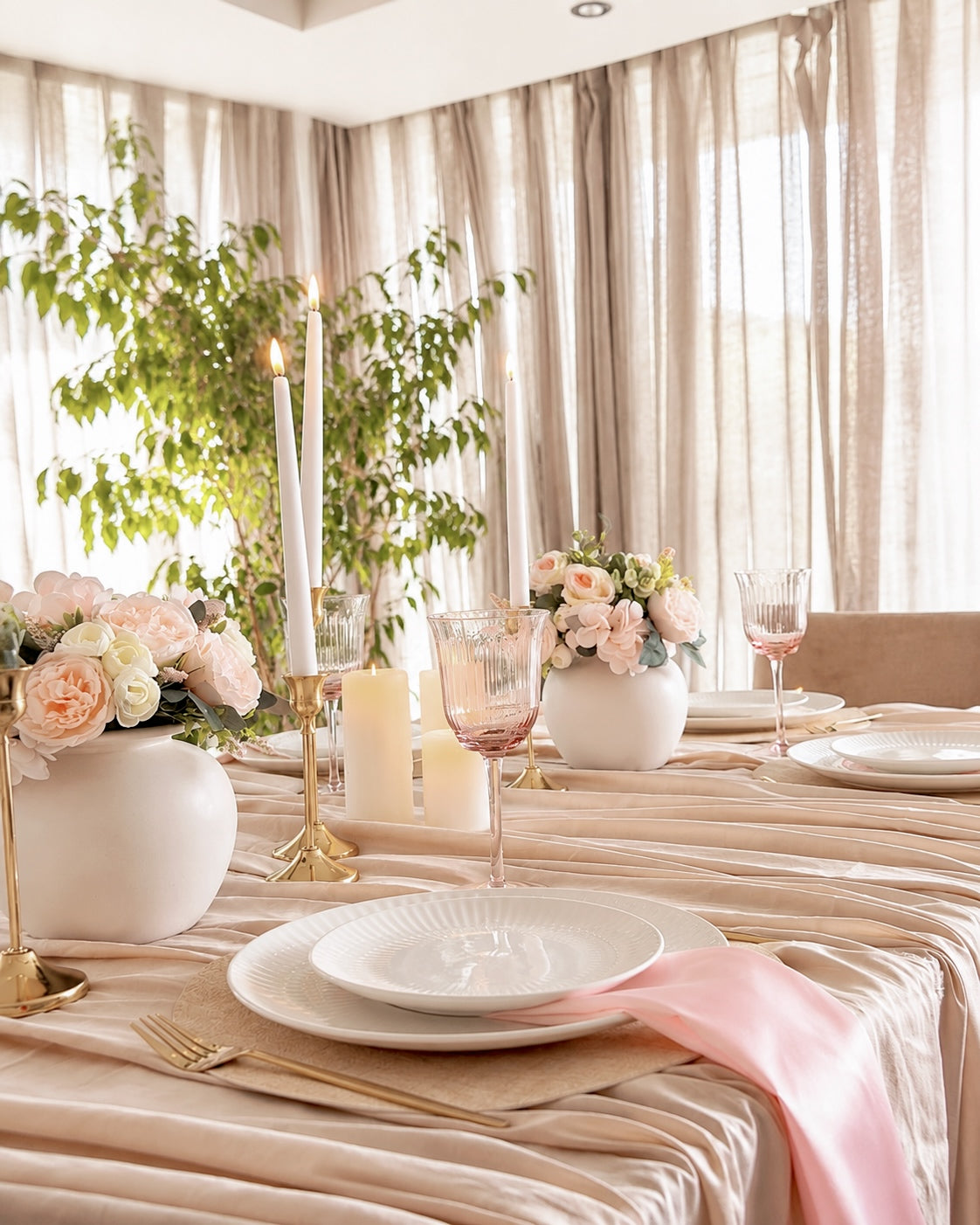 A styled dining table set in a softly lit room with beige curtains, featuring white plates, gold cutlery, pink floral arrangements, white candles, and a pale pink satin napkin.